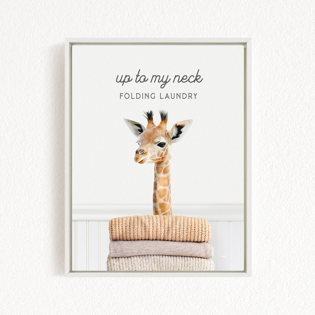 A framed image of a giraffe standing on a stack of folded towels, with the text "up to my neck" and "Folding laundry" written above it.