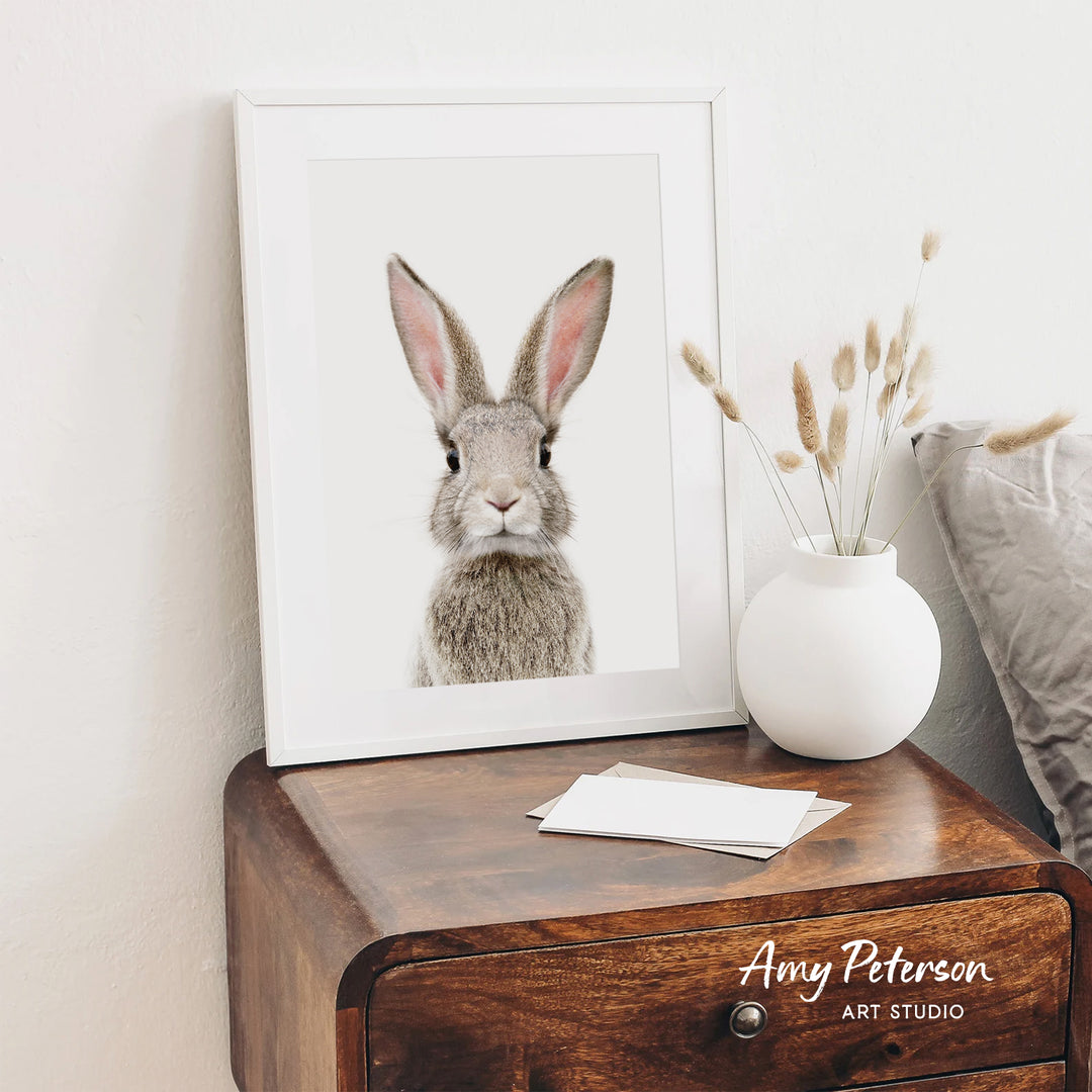 a framed picture of a rabbit on a wooden dresser, with a white vase of dried flowers and a note on the dresser.