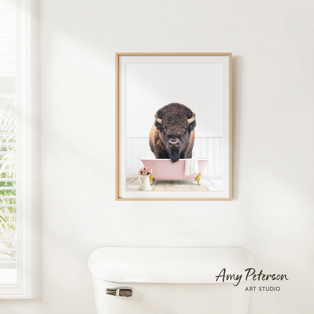 A framed photograph of a bison standing in a pink bathtub, with a white toilet and a window in the background.