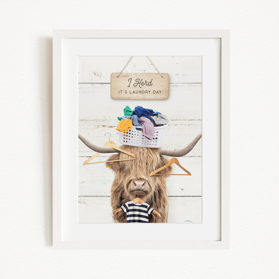A framed image depicting a cow with a basket of laundry on its head, accompanied by a sign that reads "I herd it's laundry day".