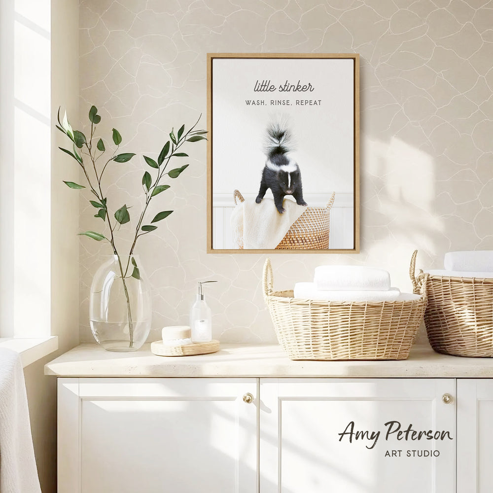 a bathroom setting with a framed poster of a skunk, a vase with green leaves, and baskets on a counter.