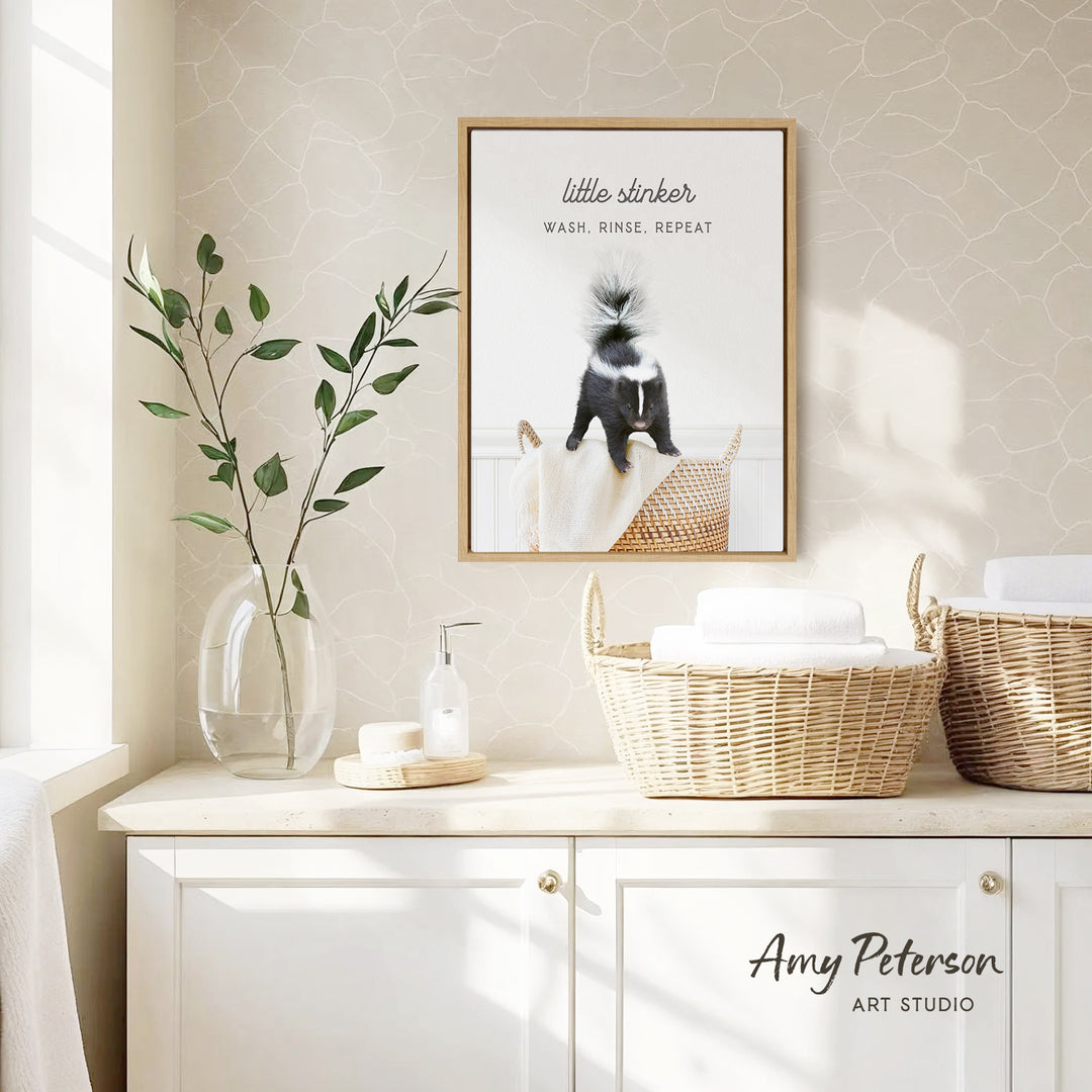 a bathroom setting with a framed poster of a skunk, a vase with green leaves, and baskets on a counter.