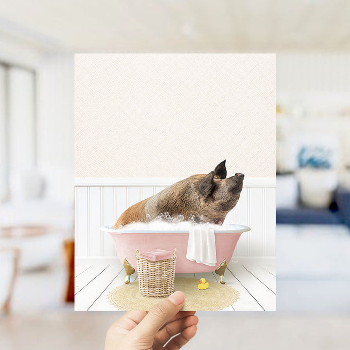 A hand holds up a framed image depicting a pig sitting in a pink bathtub filled with bubbles, surrounded by a yellow rubber duck and a white towel.