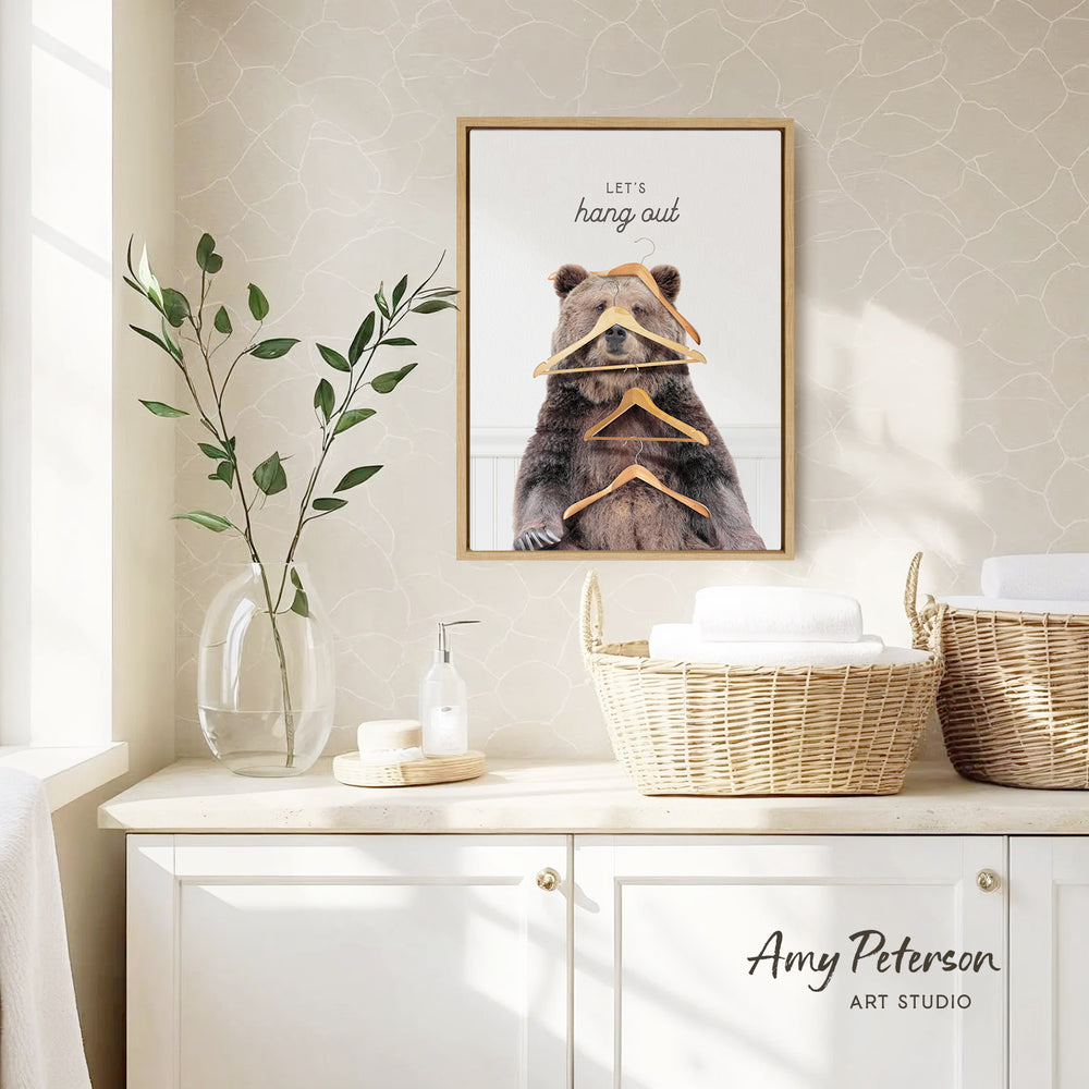 a framed poster of a bear with a humorous caption, hanging on a wall above a white cabinet with baskets and a vase of leaves.