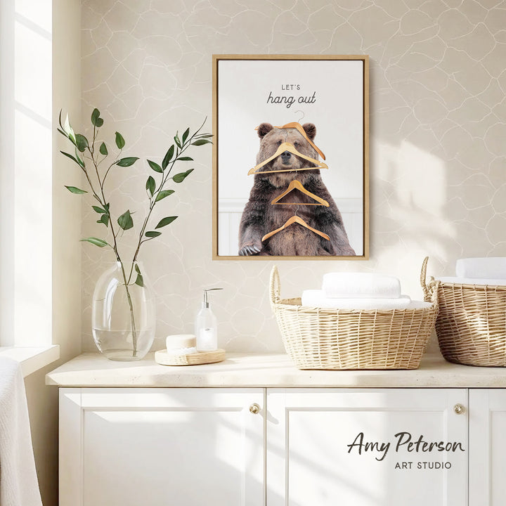 a framed poster of a bear with a humorous caption, hanging on a wall above a white cabinet with baskets and a vase of leaves.