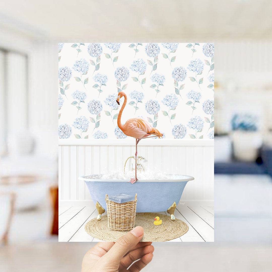 A hand holds up a framed image depicting a pink flamingo standing on a blue bathtub filled with bubbles, with a blue and white floral pattern on the wall behind it.