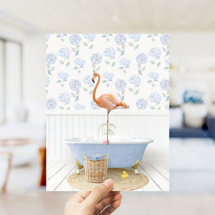 A hand holds up a framed image depicting a pink flamingo standing on a blue bathtub filled with bubbles, with a blue and white floral pattern on the wall behind it.