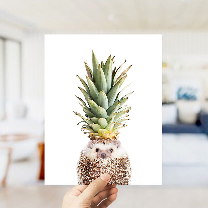 A hand holds up a photograph of a hedgehog wearing a pineapple crown.
