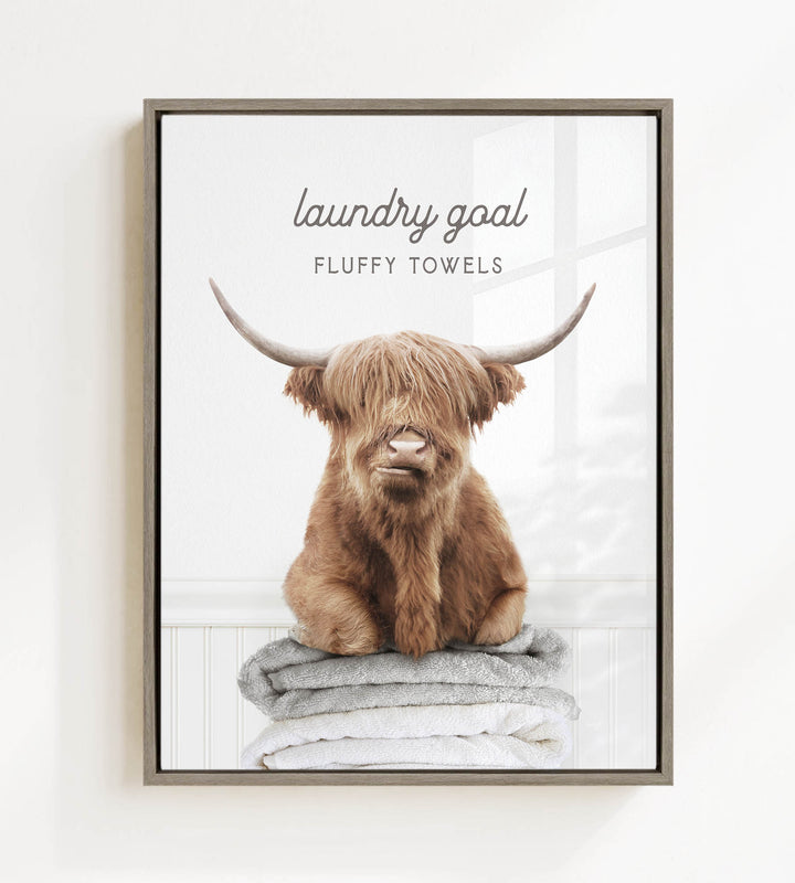 A framed image of a brown cow with large horns sitting on a stack of fluffy towels, with the text "laundry goal" and "FLUFFY TOWELS" written above it.