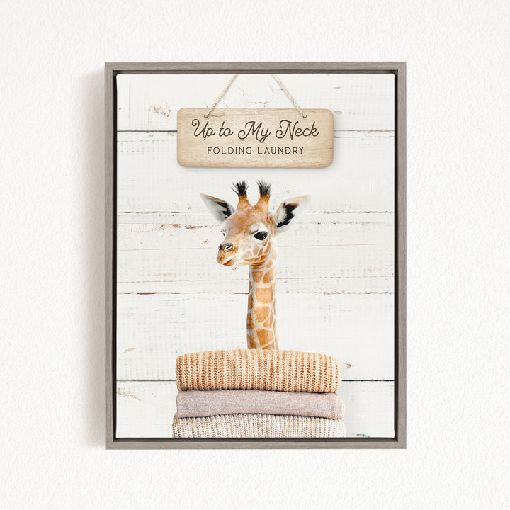 A framed image of a giraffe standing on a stack of folded towels, with a wooden sign above it that says "Up to My Neck Folding Laundry".