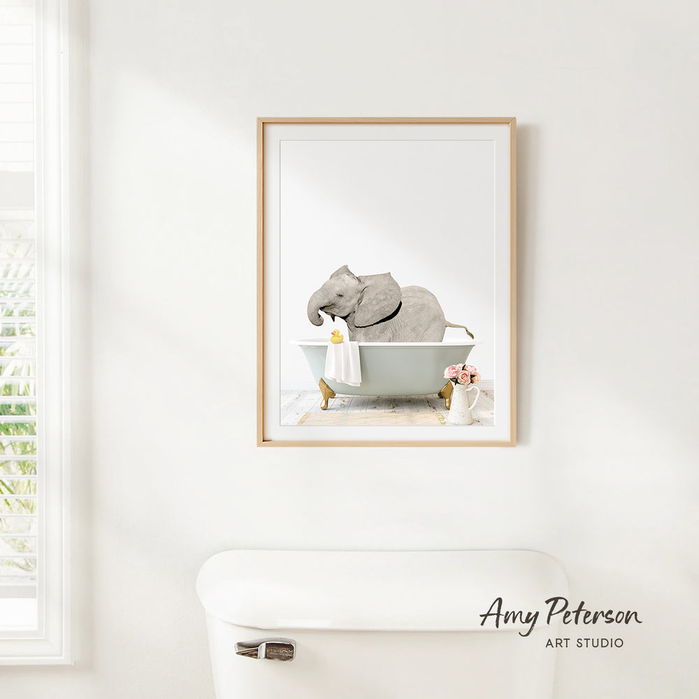 A framed print of an elephant sitting in a bathtub is hanging on a white wall in a bathroom.