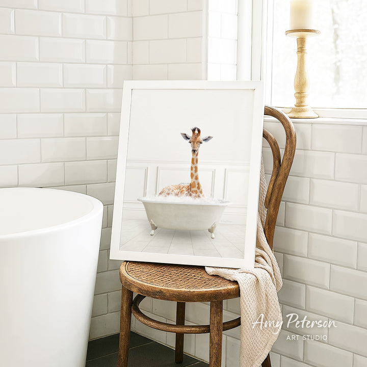 A framed image of a giraffe sitting in a bathtub, surrounded by white tiles, is displayed on a wooden chair in a bathroom setting.