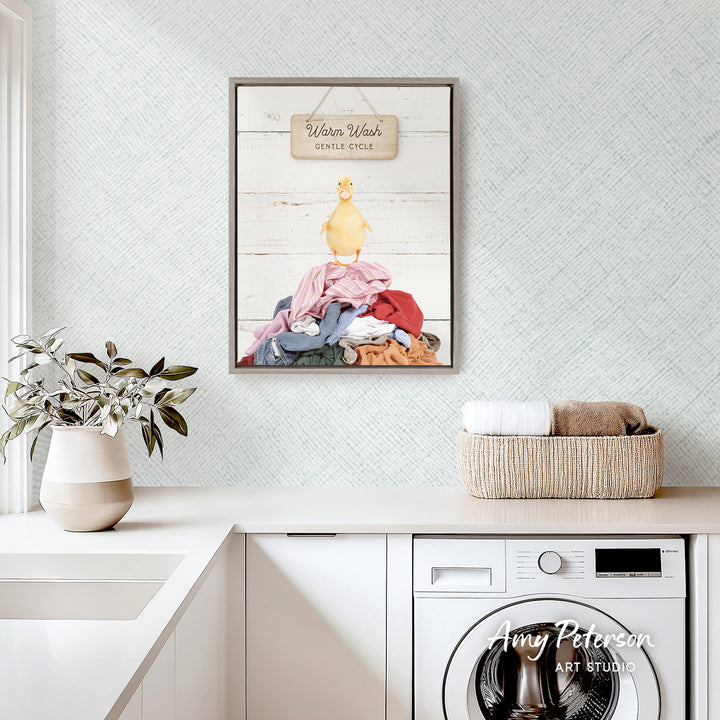 a laundry room with a framed print of a duck standing on a pile of clothes, a basket of folded towels, and a potted plant on the counter.