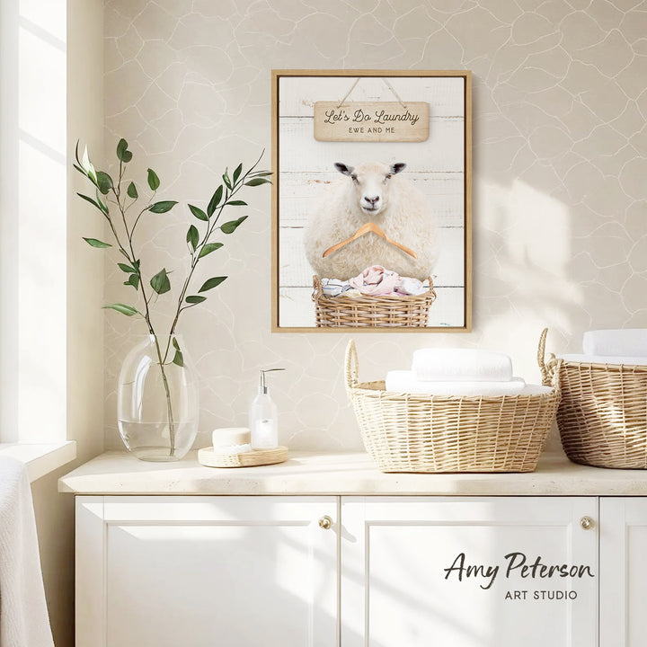 a framed picture of a sheep hanging on a wall, with a basket of towels below it. The wall has a beige color and has a window in the background. There is also a vase with a plant and a soap dispenser on a counter.