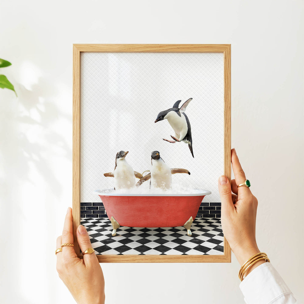 A person's hands are holding a framed picture of two penguins sitting in a red bathtub, with one penguin flying out of the tub.