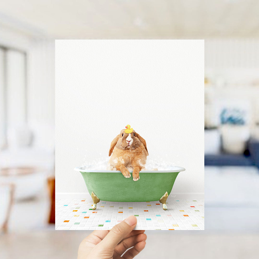 A hand holds up a framed image of a brown rabbit sitting in a green bathtub filled with bubbles, with a yellow rubber duck floating on the water.