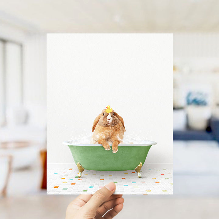 A hand holds up a framed image of a brown rabbit sitting in a green bathtub filled with bubbles, with a yellow rubber duck floating on the water.