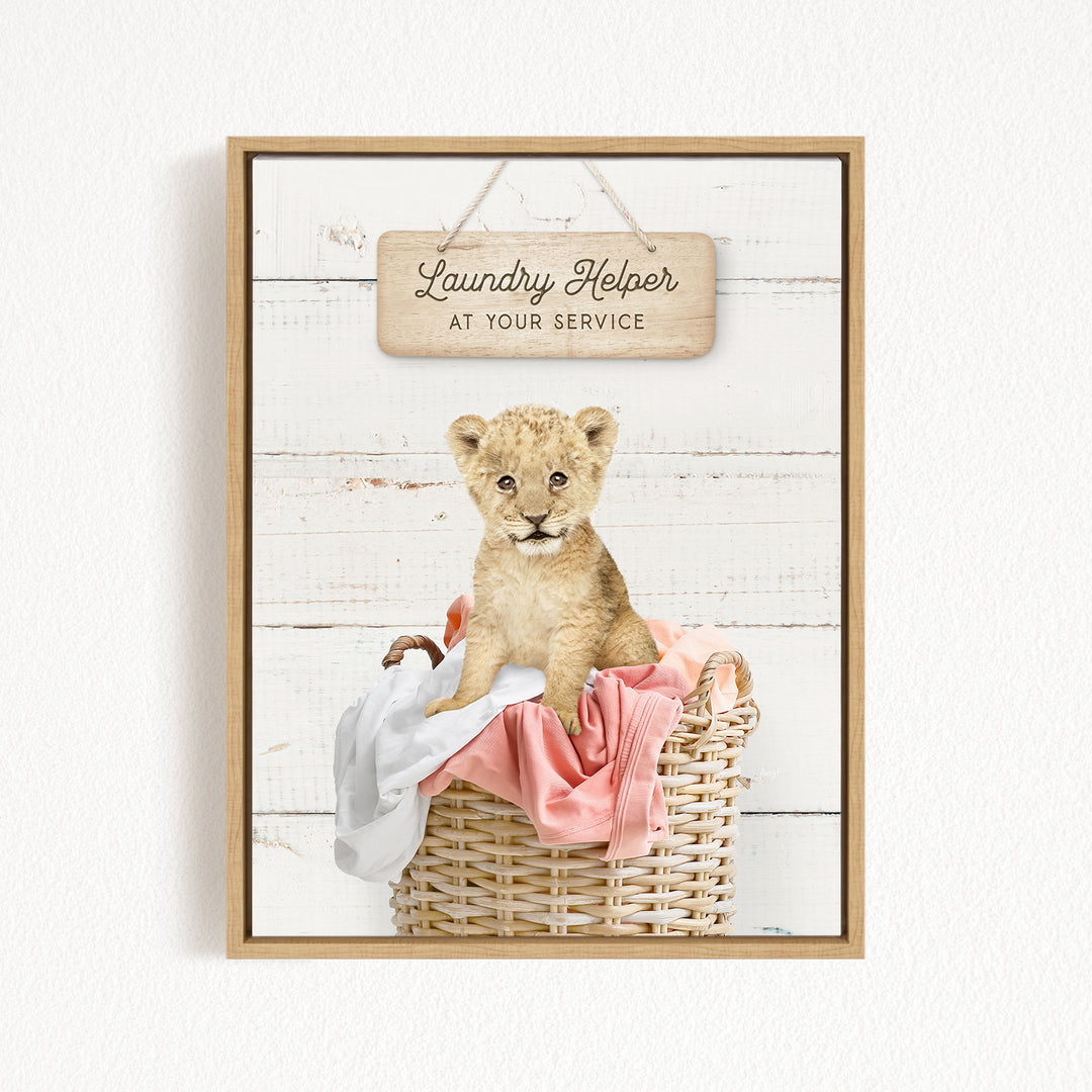 A young lion cub sitting in a wicker basket filled with clothes, with a wooden sign above that reads "Laundry Helper at your service".