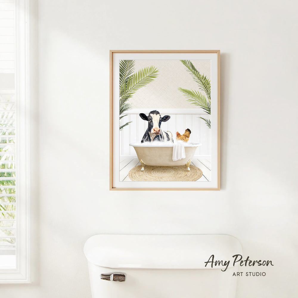 A framed print of a cow sitting in a bathtub, surrounded by palm leaves, is hanging on a white wall in a bathroom.