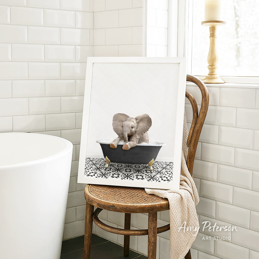 A framed image of a baby elephant sitting in a bathtub, with a towel draped over the back of a wooden chair in a bathroom setting.