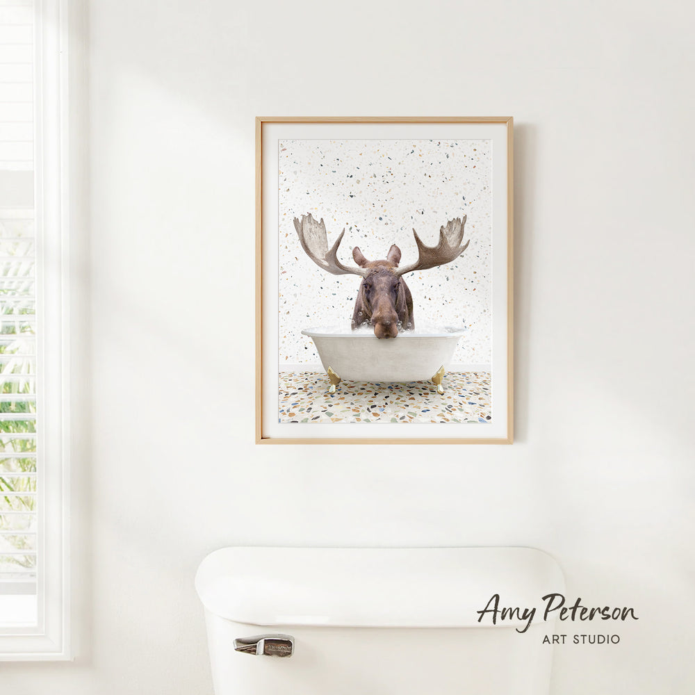 A framed print of a moose sitting in a bathtub, with its antlers spread out, is hanging on a white wall.