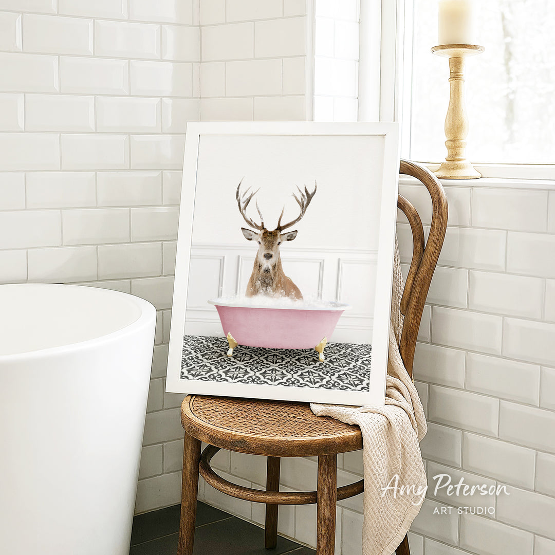 A framed image of a deer with antlers sitting in a pink bathtub, surrounded by a patterned rug, is displayed on a wooden chair in a bathroom setting.
