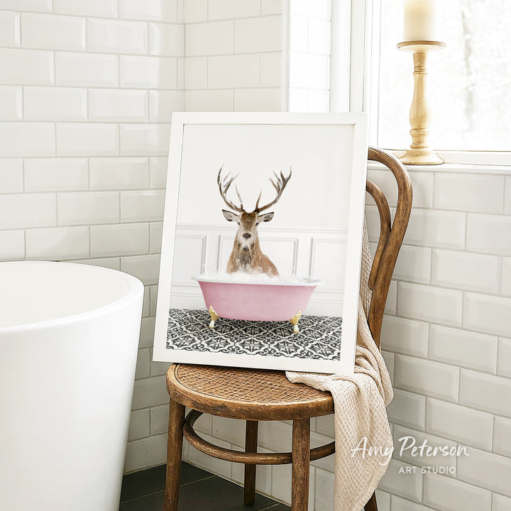 A framed image of a deer with antlers sitting in a pink bathtub, surrounded by a patterned rug, is displayed on a wooden chair in a bathroom setting.