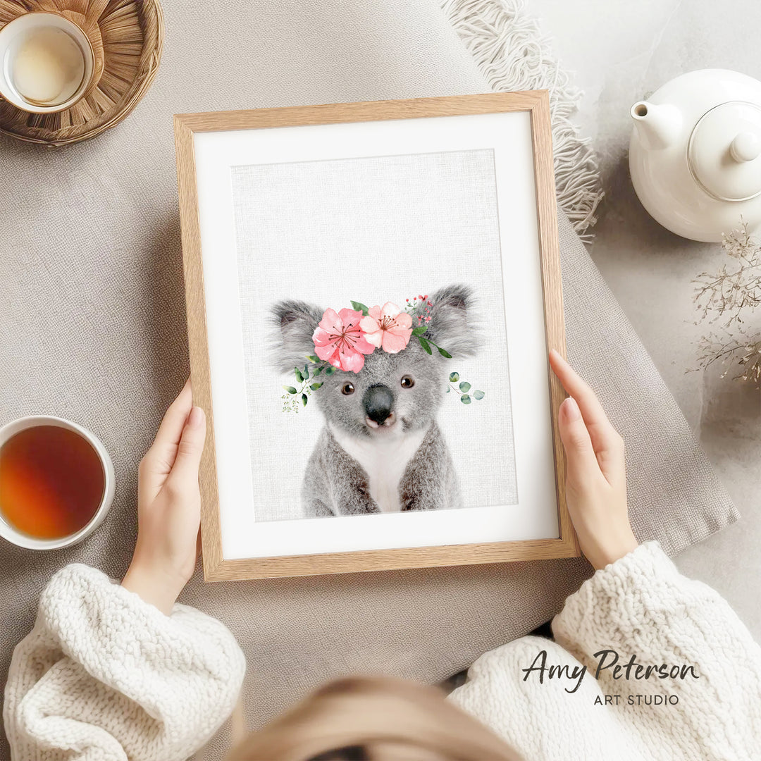 A person is holding a framed picture of a koala bear wearing a flower crown, with a teapot and a cup of tea nearby.
