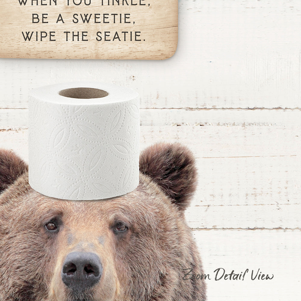 A brown bear with a roll of toilet paper on its head, with a wooden sign above it that says "When you tinkle, be a sweetie, wipe the seatie."