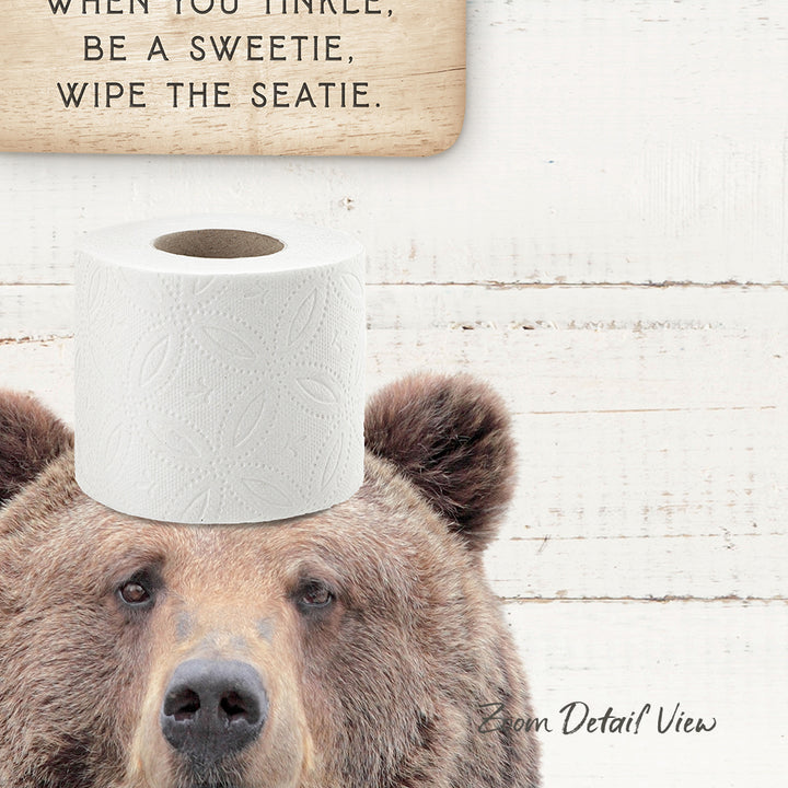 A brown bear with a roll of toilet paper on its head, with a wooden sign above it that says "When you tinkle, be a sweetie, wipe the seatie."