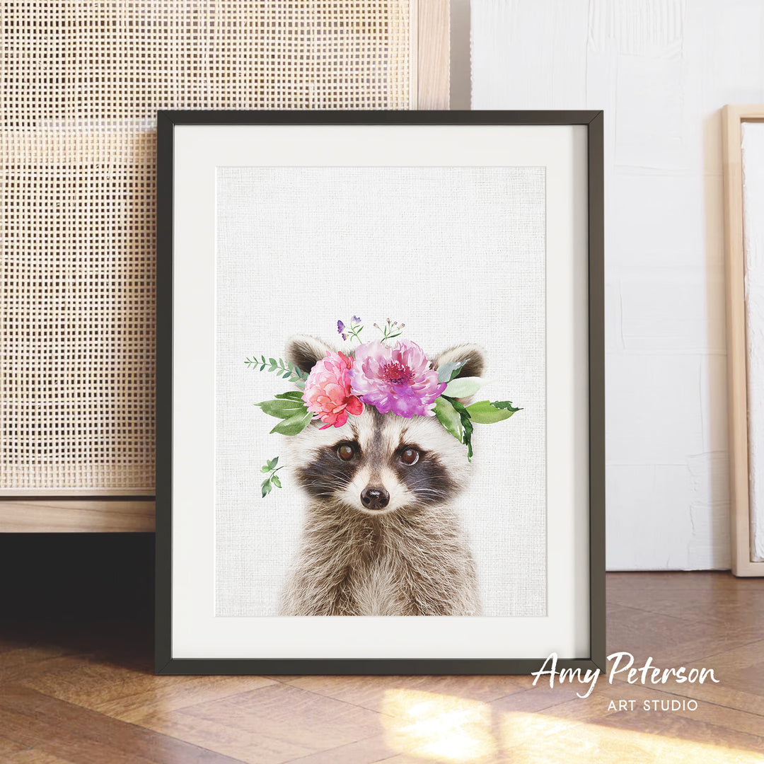 A framed print of a raccoon wearing a floral headpiece is displayed on a wooden floor against a white wall.