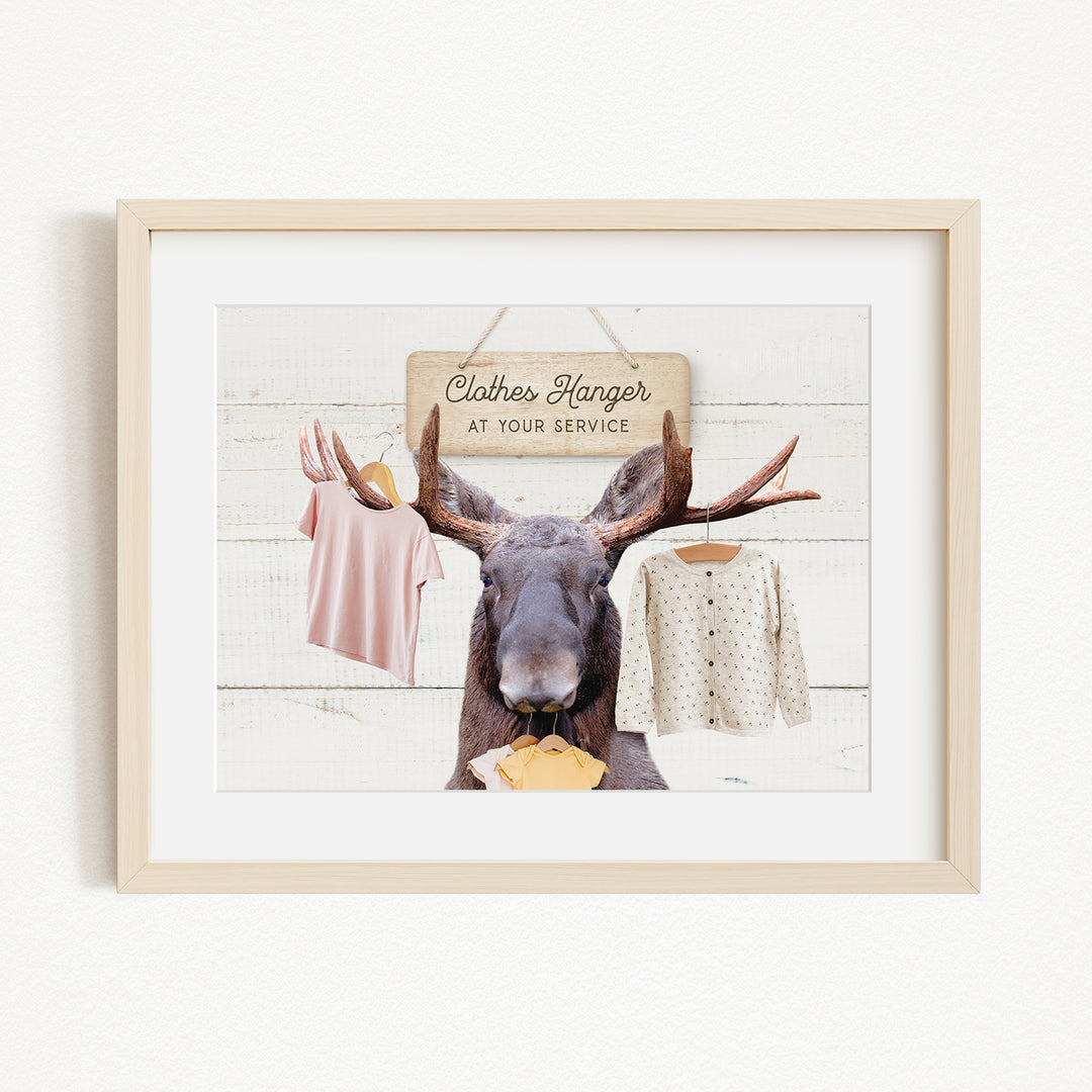 A framed image depicting a moose with antlers holding clothes, including a pink shirt and a white sweater, against a white background with a wooden sign that reads "Clothes Hanger at your service".