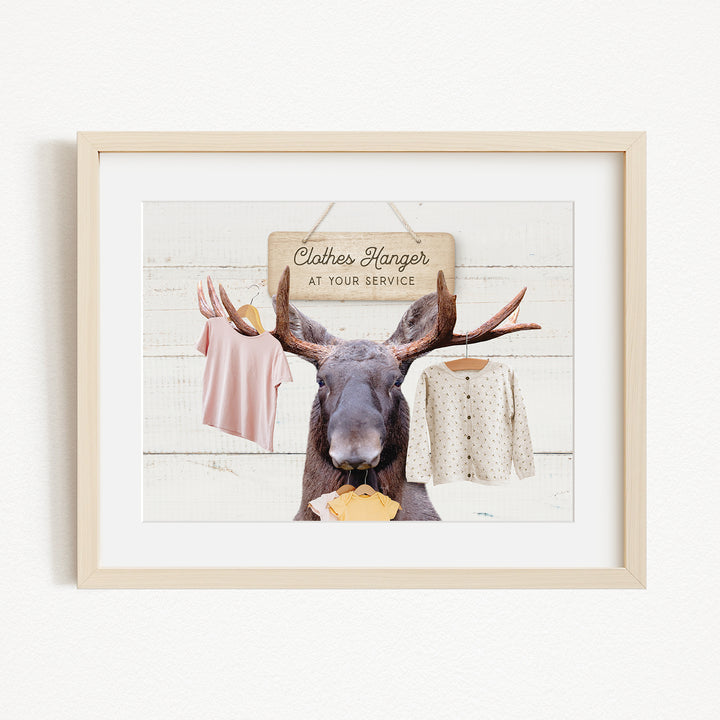 A framed image depicting a moose with antlers holding clothes, including a pink shirt and a white sweater, against a white background with a wooden sign that reads "Clothes Hanger at your service".
