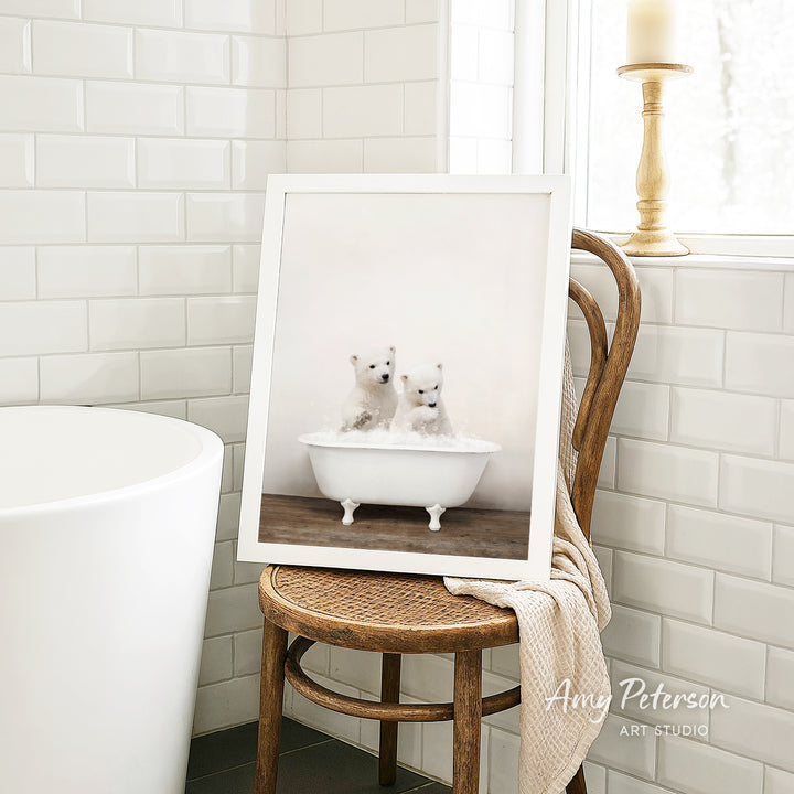 A framed photograph of two adorable polar bears sitting in a white bathtub, with a wooden chair and a white tiled wall in the background.