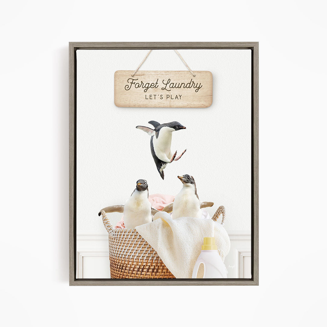A framed image depicting three penguins in a basket, with a sign above that says "Forget Laundry, Let's Play".