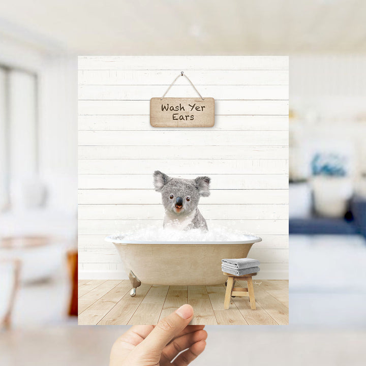 A hand holds up a framed image of a koala sitting in a bathtub, with a sign above that reads "Wash Your Ears".