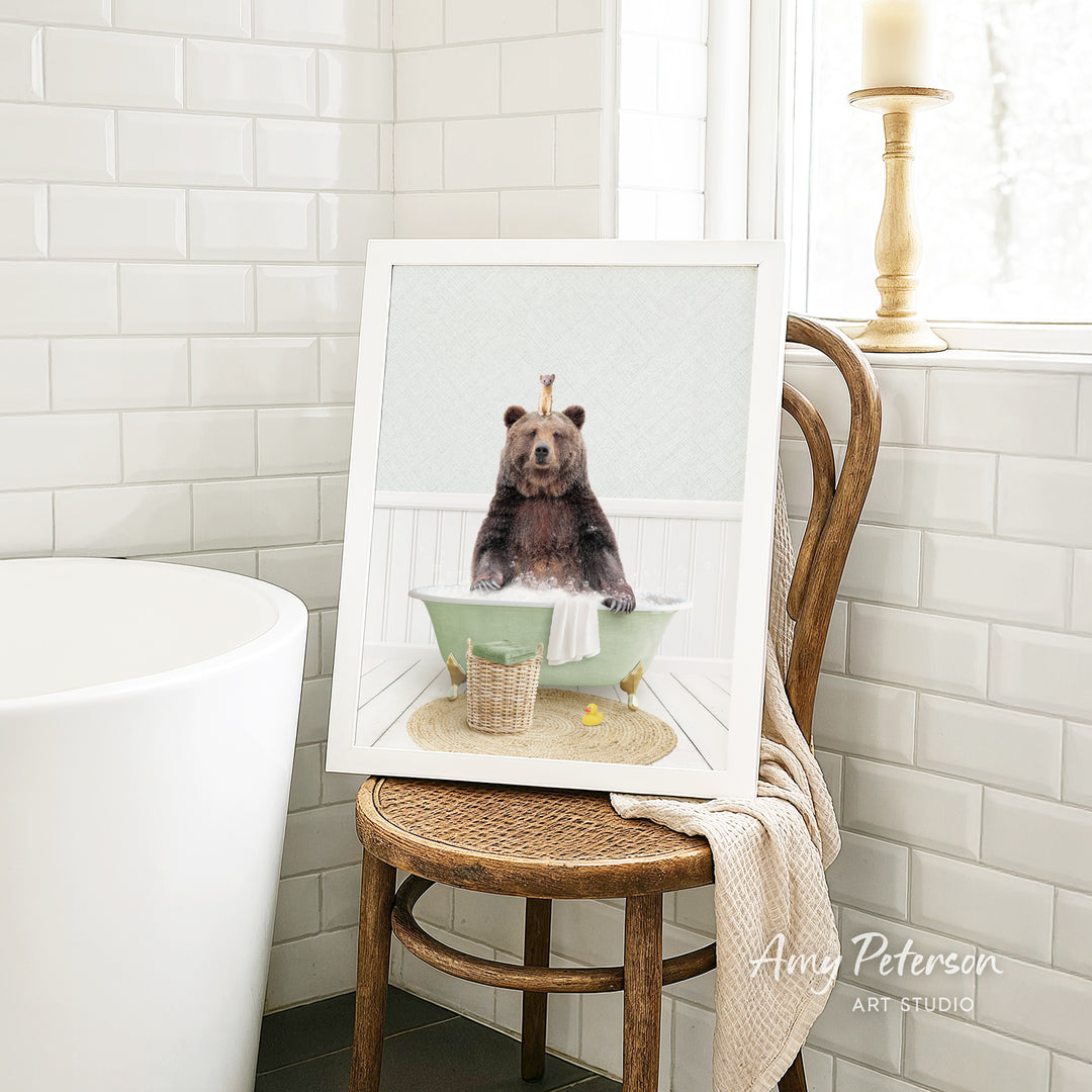 A framed image of a bear sitting in a bathtub, with a towel and basket nearby, is displayed on a wooden chair in a bathroom setting.