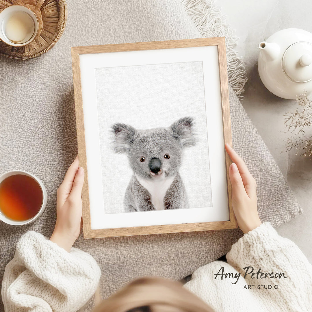 A person is holding a framed picture of a koala bear, which is the central focus of the image. The background includes a teapot, a cup, and a bowl, suggesting a cozy and relaxed setting.