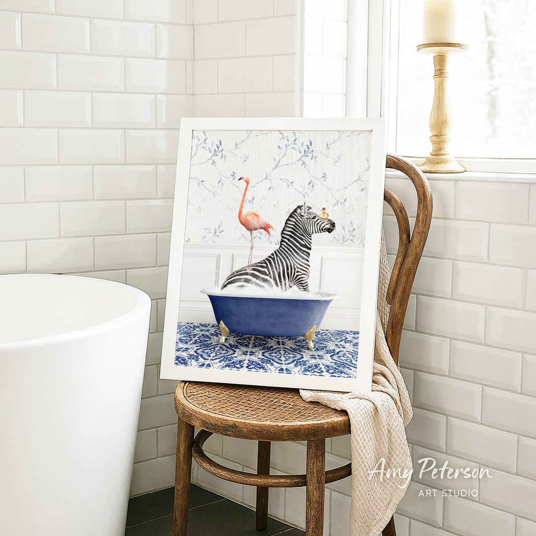 A framed painting of a zebra sitting in a blue bathtub, with a pink flamingo standing on its back, is displayed on a wooden chair in a bathroom setting.