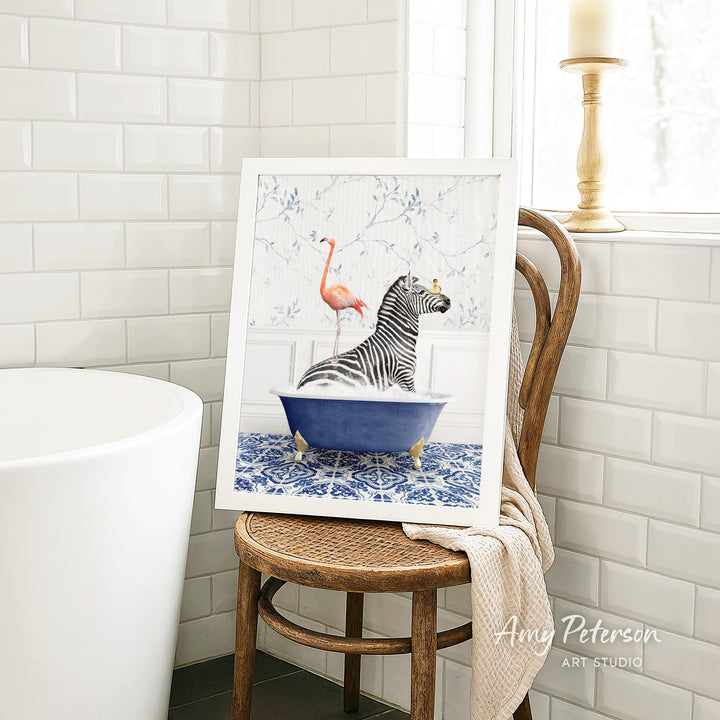 A framed painting of a zebra sitting in a blue bathtub, with a pink flamingo standing on its back, is displayed on a wooden chair in a bathroom setting.