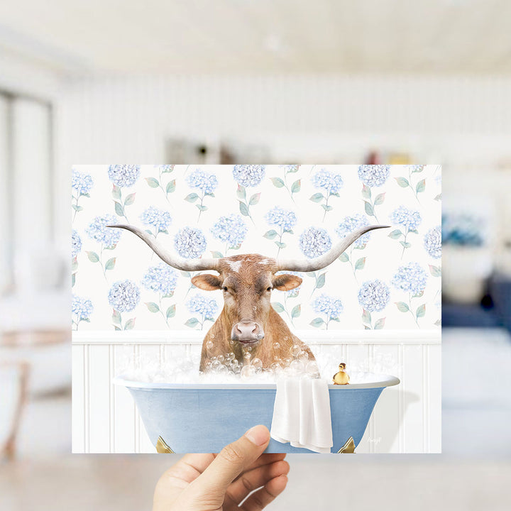 A brown cow with large horns is sitting in a blue bathtub filled with water, surrounded by a floral pattern.