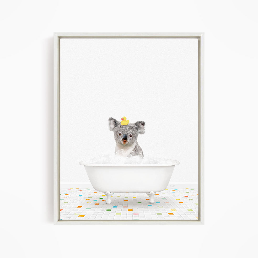 A koala bear sitting in a white bathtub filled with bubbles, with a rubber duck perched on its head.
