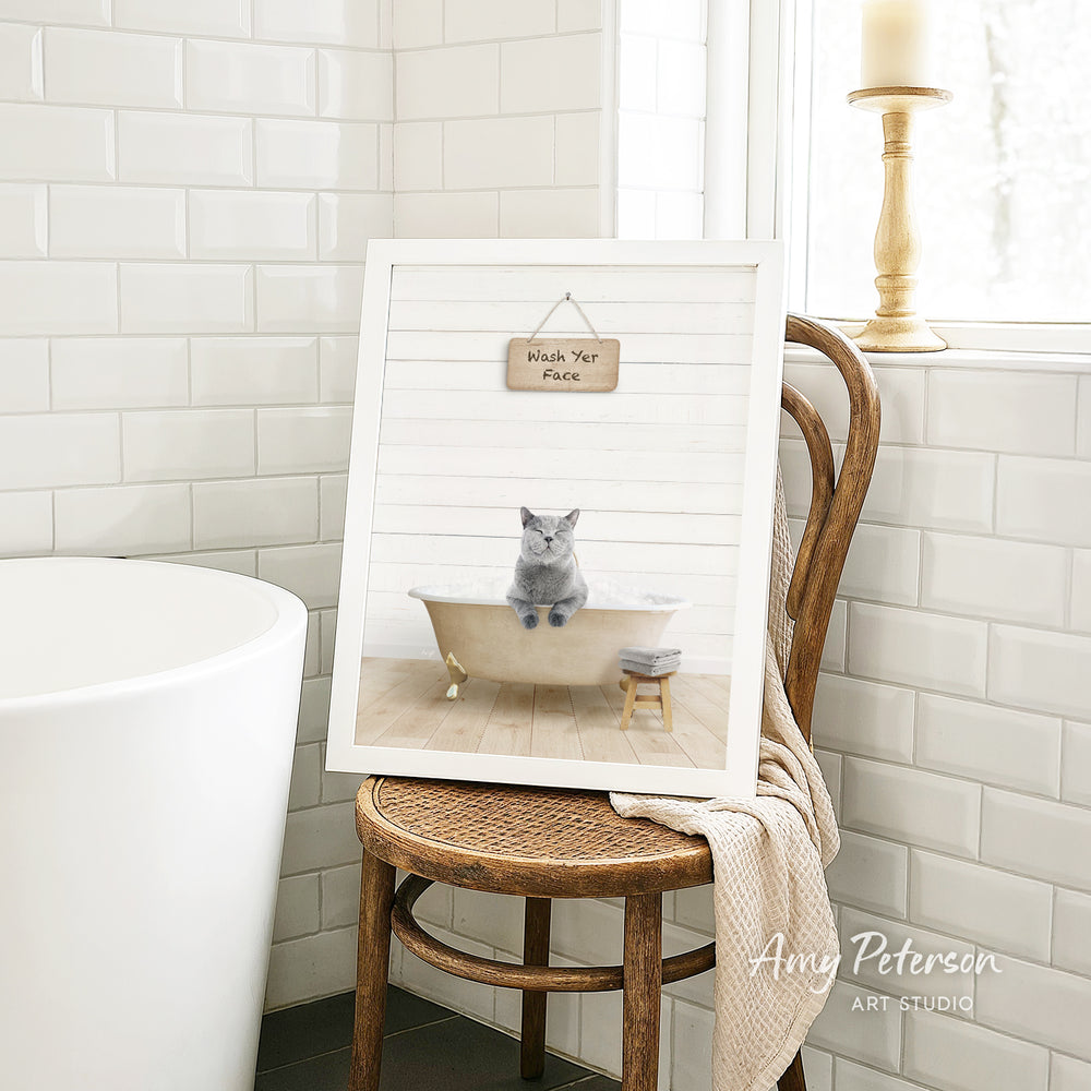 A framed image of a cat sitting in a bathtub, with a sign above it that says "Wash Your Face". The image is displayed on a wooden chair in a bathroom setting.