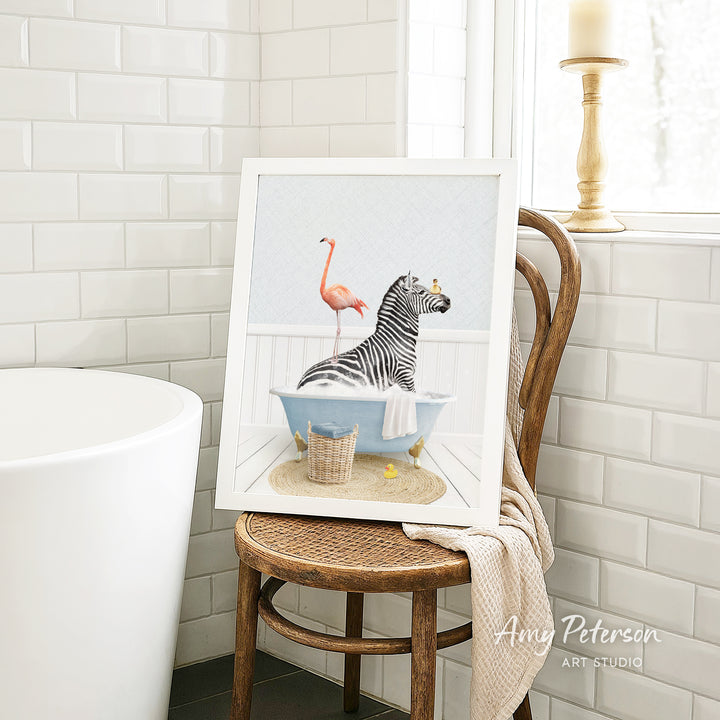 A framed painting of a zebra sitting in a bathtub, with a pink flamingo standing on its back, is displayed on a wooden chair in a bathroom setting.