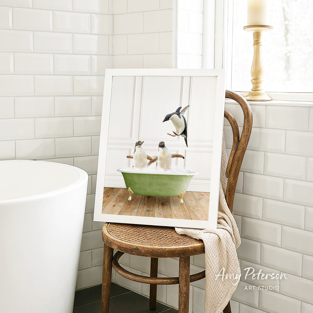 A framed image of penguins sitting in a bathtub is displayed on a wooden chair in a bathroom setting.