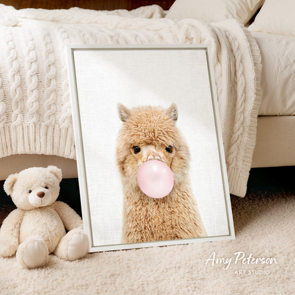 A framed image of a fluffy alpaca blowing a bubble with a pink bubble gum bubble, accompanied by a teddy bear sitting next to it.