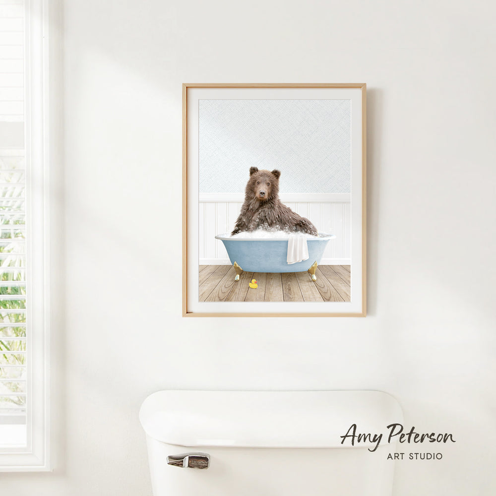 A framed image of a brown bear sitting in a blue bathtub, with a towel draped over the side of the tub.