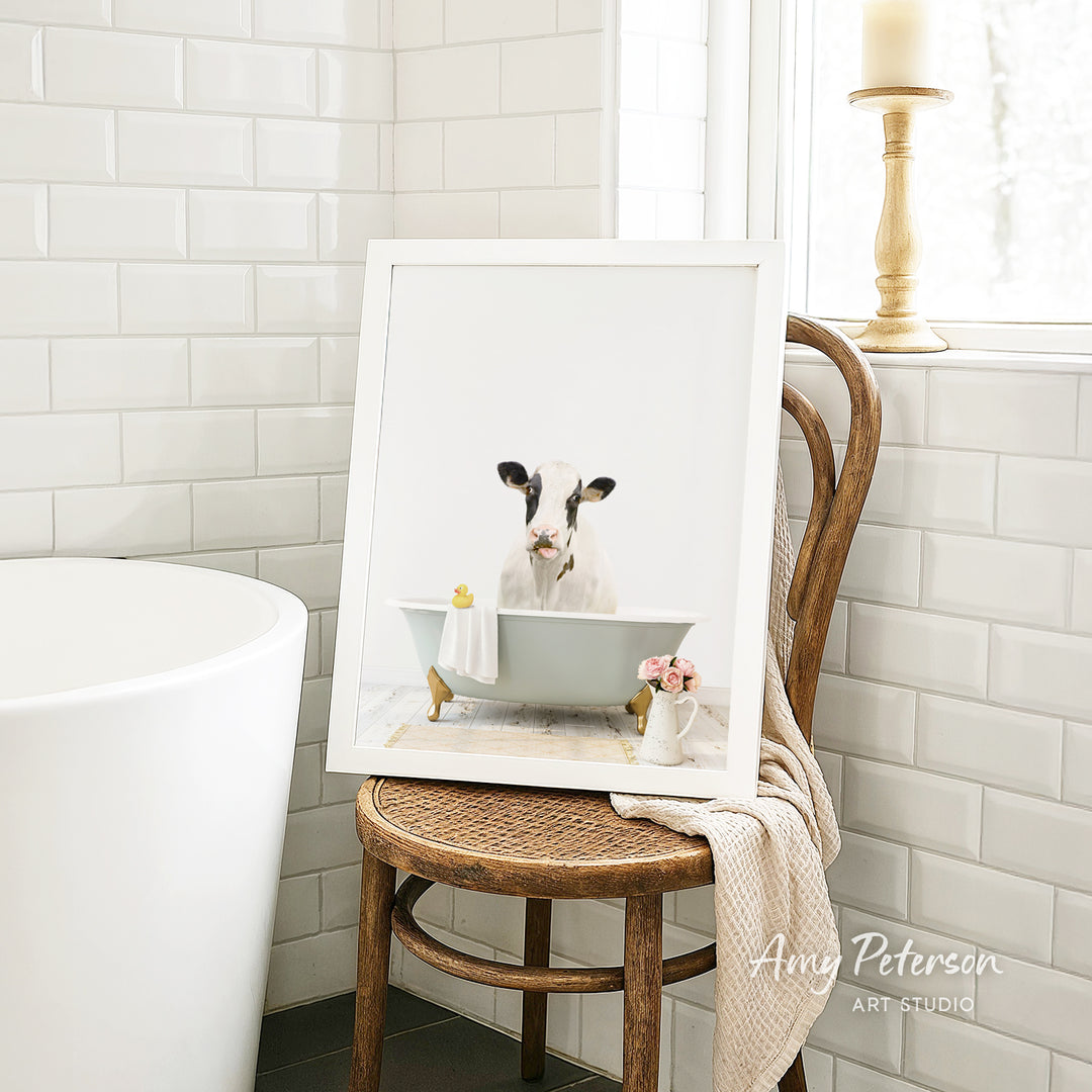 A framed image of a cow sitting in a bathtub, with a vase of flowers and a candle on a wooden chair in the background.