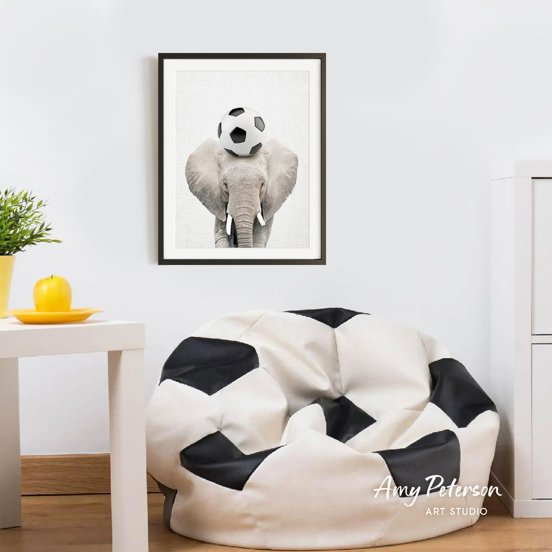 a framed picture of an elephant with a soccer ball on its head, hanging on a white wall above a white table with a yellow plate on it.