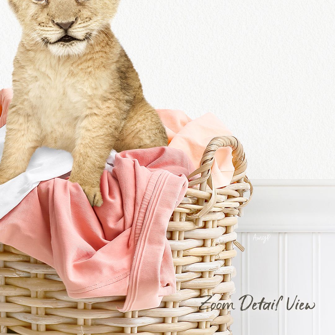 A young lion cub sitting in a wicker basket filled with pink and white clothing.